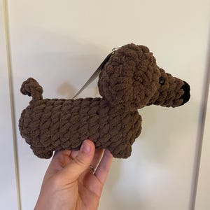 May include: A brown crocheted dachshund plush toy with a black nose and a small curled tail. The stuffed animal is crafted from thick, textured yarn and features floppy ears, a long body, and a small paper tag attached to the top of its head.