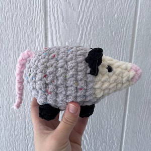 May include: A handmade crochet plush toy shaped like an opossum. The body is made of grey yarn with small colourful speckles, while the face is cream-coloured with a pink nose. It features black ears, black feet, and a long, textured pink tail.