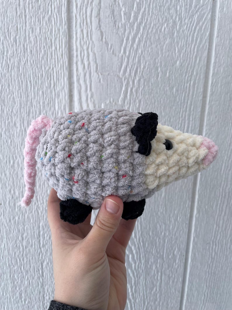 May include: A handmade crochet plush toy shaped like an opossum. The body is made of grey yarn with small colourful speckles, while the face is cream-coloured with a pink nose. It features black ears, black feet, and a long, textured pink tail.