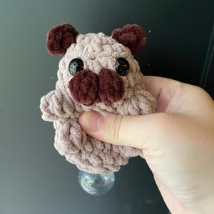 May include: A handmade, plush pug-shaped bottle stopper. The pug has a light brown body, dark brown ears and nose, and large black eyes. The stopper is held in a hand and has a clear, round glass base.