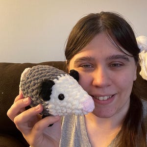 May include: A small, handmade crochet plush toy shaped like an opossum. The body is crafted from textured grey yarn, featuring a white face, small black ears, and a soft pink nose. It has two small, round, black plastic eyes set into the white yarn.