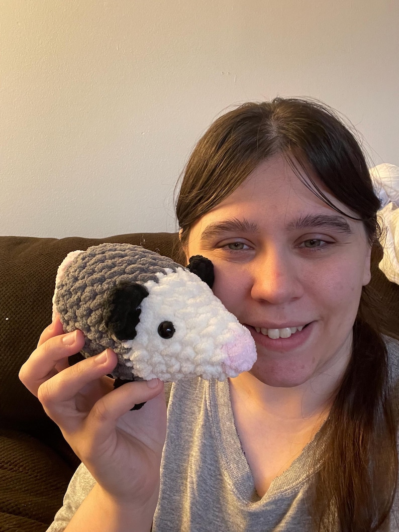 May include: A small, handmade crochet plush toy shaped like an opossum. The body is crafted from textured grey yarn, featuring a white face, small black ears, and a soft pink nose. It has two small, round, black plastic eyes set into the white yarn.