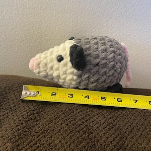 May include: A crocheted plush opossum with a white face, pink nose, black ears, and a grey body with a pink tail. A yellow measuring tape is positioned along the base, showing the plush measures approximately 17.8 cm in total length.