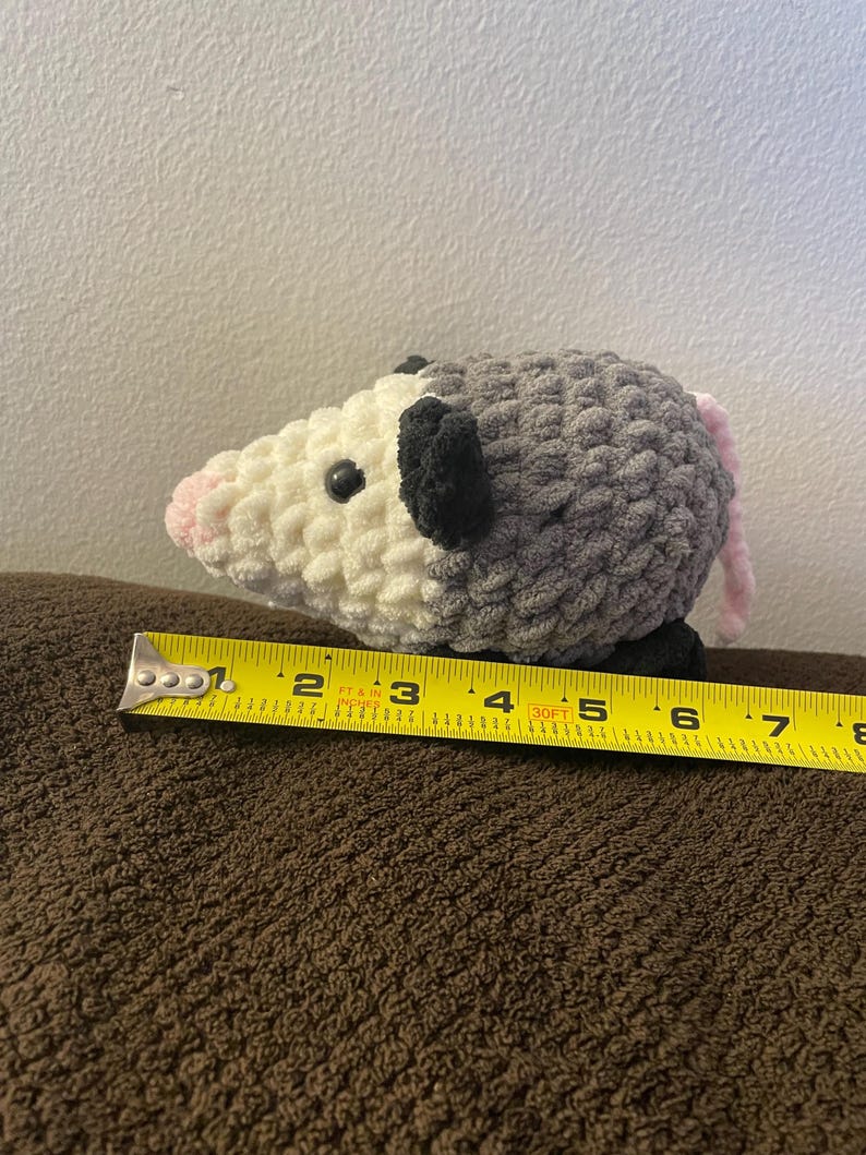May include: A crocheted plush opossum with a white face, pink nose, black ears, and a grey body with a pink tail. A yellow measuring tape is positioned along the base, showing the plush measures approximately 17.8 cm in total length.