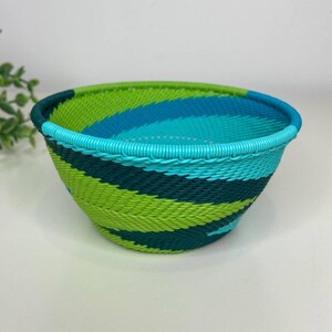 May include: A woven bowl with a vibrant color scheme. The bowl features a combination of green, teal, and blue hues in a striped pattern. The rim of the bowl is a solid teal color. The bowl is likely used for storage or decoration.