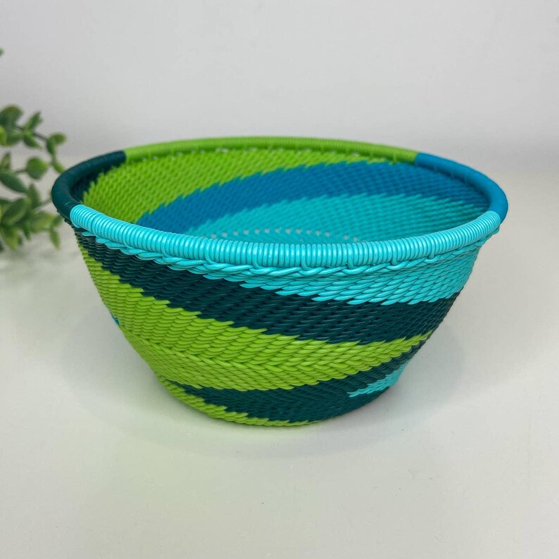 May include: A woven bowl with a vibrant color scheme. The bowl features a combination of green, teal, and blue hues in a striped pattern. The rim of the bowl is a solid teal color. The bowl is likely used for storage or decoration.