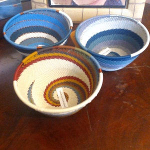 May include: Three woven baskets with spiral designs. The baskets are in shades of blue, white, red, and yellow. The baskets are round and have a textured appearance. The baskets are sitting on a dark brown wooden surface.