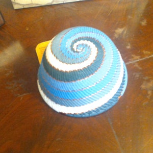 May include: A woven basket with a spiral design in shades of blue, white, and dark blue. The basket has a rounded shape and appears to be handmade. The colors create a visually appealing pattern, suitable for home decor or storage.