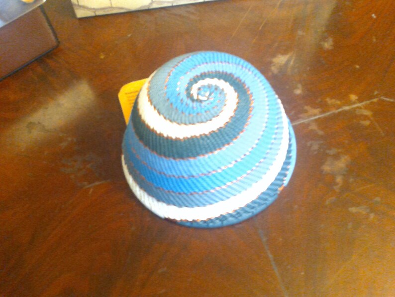May include: A woven basket with a spiral design in shades of blue, white, and dark blue. The basket has a rounded shape and appears to be handmade. The colors create a visually appealing pattern, suitable for home decor or storage.