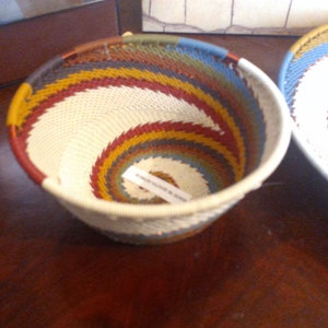 May include: A handwoven basket with a spiral design. The basket features a cream-colored center with bands of red, yellow, blue, and green. The rim is cream-colored. A second basket is partially visible in the background.