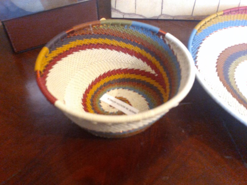 May include: A handwoven basket with a spiral design. The basket features a cream-colored center with bands of red, yellow, blue, and green. The rim is cream-colored. A second basket is partially visible in the background.