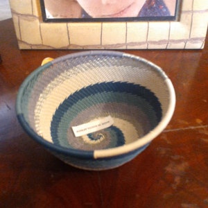 May include: Handwoven basket with a spiral design in shades of blue, gray, and cream. The basket has a rounded shape and a sturdy rim. A small tag is attached to the basket. The basket is sitting on a wooden surface.