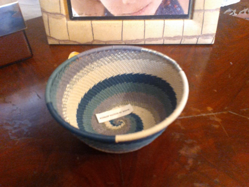 May include: Handwoven basket with a spiral design in shades of blue, gray, and cream. The basket has a rounded shape and a sturdy rim. A small tag is attached to the basket. The basket is sitting on a wooden surface.