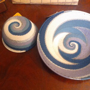 May include: Two woven baskets with a spiral design in shades of blue, white, and gray. One is a bowl shape, and the other is a smaller, rounded shape. The baskets are placed on a dark wooden surface.