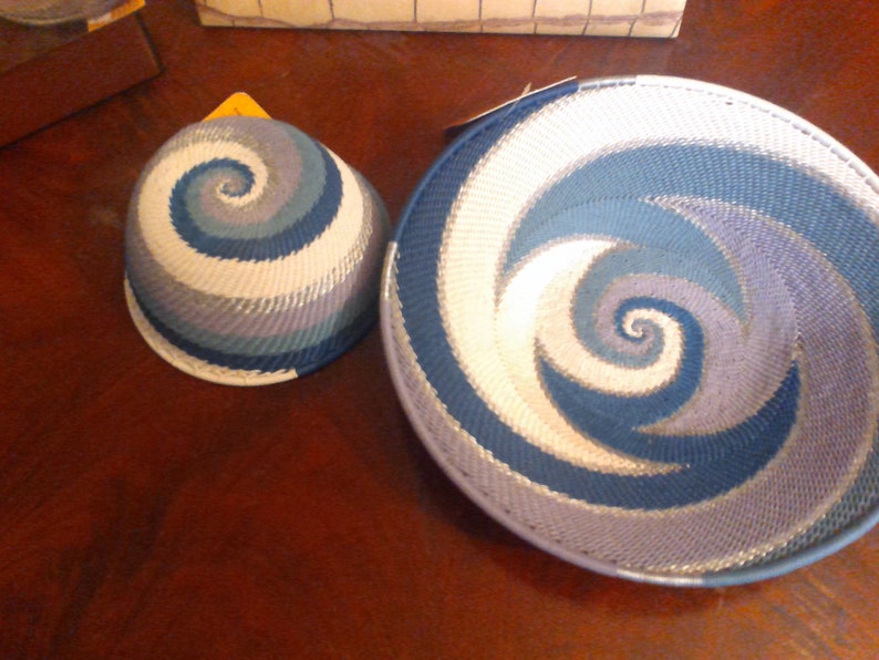 May include: Two woven baskets with a spiral design in shades of blue, white, and gray. One is a bowl shape, and the other is a smaller, rounded shape. The baskets are placed on a dark wooden surface.
