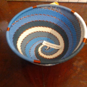 May include: A woven basket with a spiral design in shades of blue, white, and gray. The basket has a rounded shape with a blue rim and orange accents. The text on the label reads "MADE IN SOUTH AFRICA".