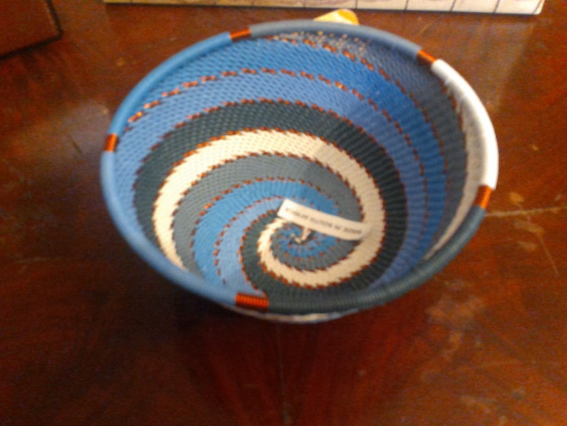 May include: A woven basket with a spiral design in shades of blue, white, and gray. The basket has a rounded shape with a blue rim and orange accents. The text on the label reads "MADE IN SOUTH AFRICA".