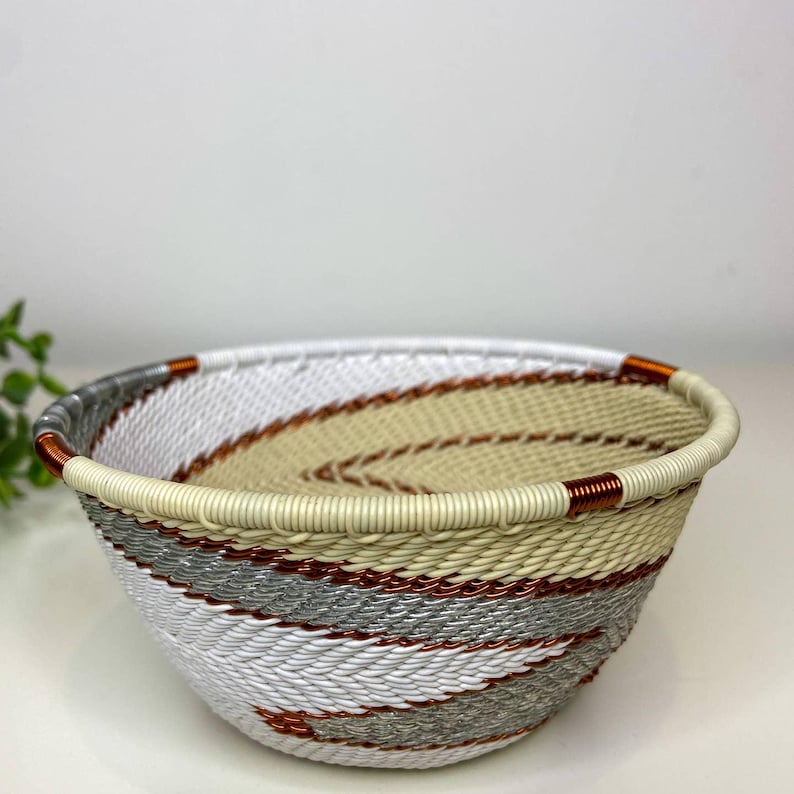 May include: A woven bowl with a cream-colored rim and a spiral design. The bowl features alternating bands of white, gray, and tan, accented with copper-colored wire. This decorative bowl is ideal for home decor.