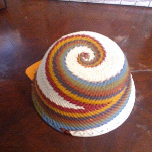 May include: A handwoven basket with a spiral design. The basket features a white base with a colorful spiral pattern in shades of red, yellow, blue, and green. The basket is likely used for storage or decoration.