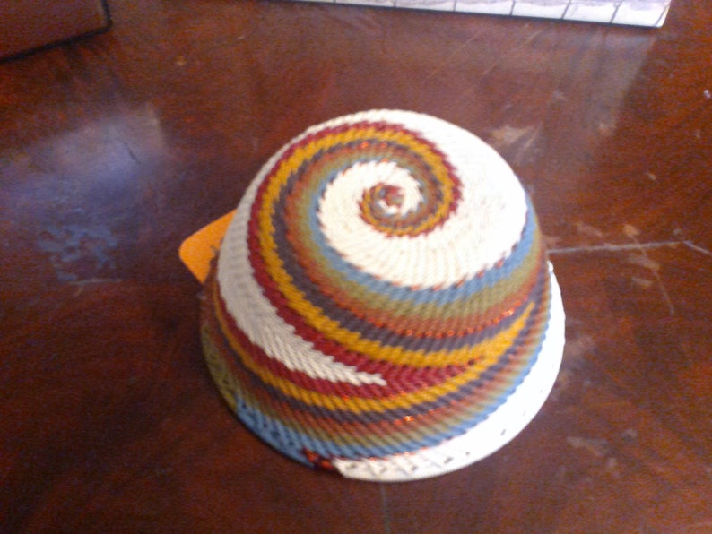 May include: A handwoven basket with a spiral design. The basket features a white base with a colorful spiral pattern in shades of red, yellow, blue, and green. The basket is likely used for storage or decoration.