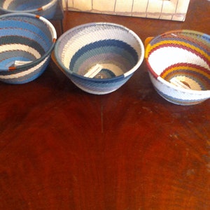 May include: Three woven bowls with colorful striped patterns. The bowls are round with a flared shape, featuring shades of blue, white, and other colors. The bowls are displayed on a dark wooden surface.