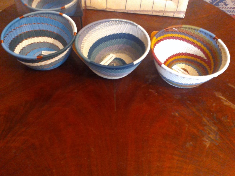 May include: Three woven bowls with colorful striped patterns. The bowls are round with a flared shape, featuring shades of blue, white, and other colors. The bowls are displayed on a dark wooden surface.