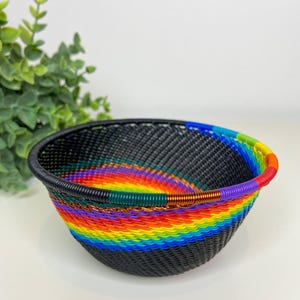 May include: A woven basket with a black base and a vibrant rainbow pattern around the middle. The rim of the basket is black, and the rainbow colors include red, orange, yellow, green, blue, and purple. The basket is likely handmade.