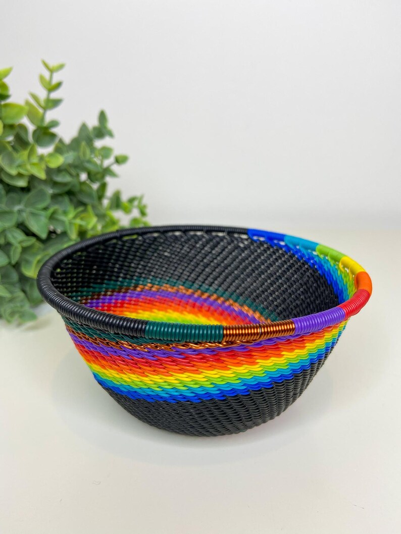 May include: A woven basket with a black base and a vibrant rainbow pattern around the middle. The rim of the basket is black, and the rainbow colors include red, orange, yellow, green, blue, and purple. The basket is likely handmade.