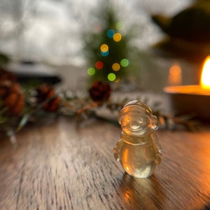 May include: A clear glass snowman figurine with a white hat and scarf. The figurine is sitting on a wooden surface with a blurred background of a Christmas tree with lights.