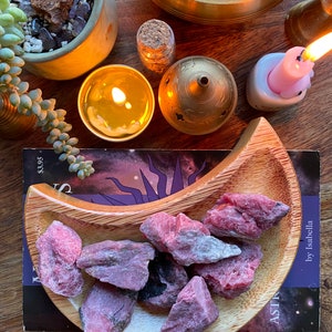 May include: A wooden crescent moon-shaped dish filled with pink and gray rhodochrosite crystals. The dish is on a book with a purple and black galaxy design. The book title is "Astro" by Isabella.