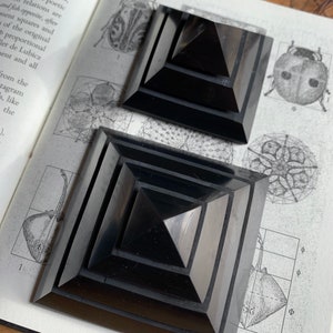 May include: Two black obsidian pyramids, one stacked on top of the other, with a geometric pattern on the base. The pyramids are on a book with a black and white illustration of a geometric pattern.
