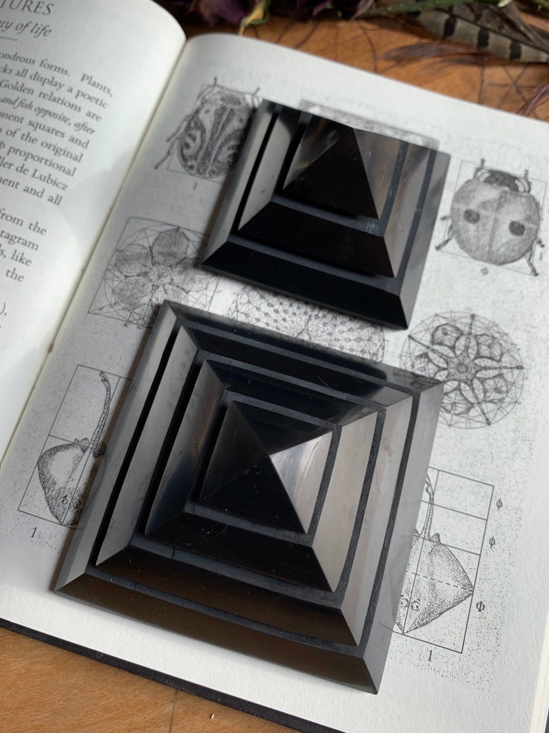 May include: Two black obsidian pyramids, one stacked on top of the other, with a geometric pattern on the base. The pyramids are on a book with a black and white illustration of a geometric pattern.