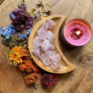 May include: A wooden crescent moon shaped dish filled with rose quartz crystals. The dish is on a wooden surface with dried flowers and a pink candle.