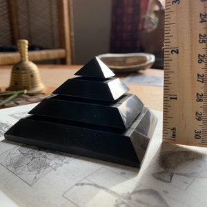 May include: A black obsidian pyramid with five tiers, measuring approximately 2 inches in height. The pyramid is resting on a book with geometric patterns.