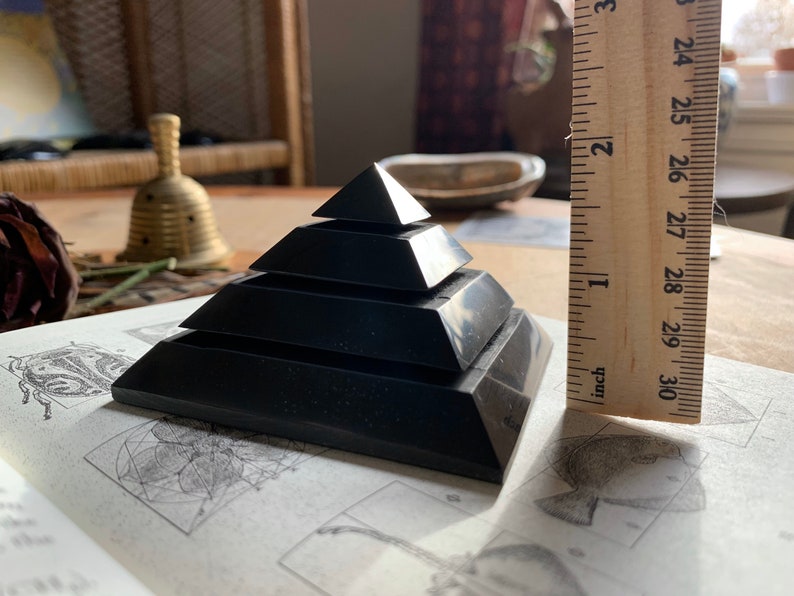 May include: A black obsidian pyramid with five tiers, measuring approximately 2 inches in height. The pyramid is resting on a book with geometric patterns.