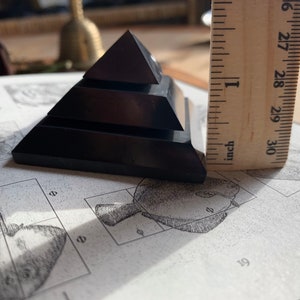 May include: A black obsidian pyramid with four tiers, measuring approximately 1 inch in height. The pyramid is resting on a page of a book with illustrations.