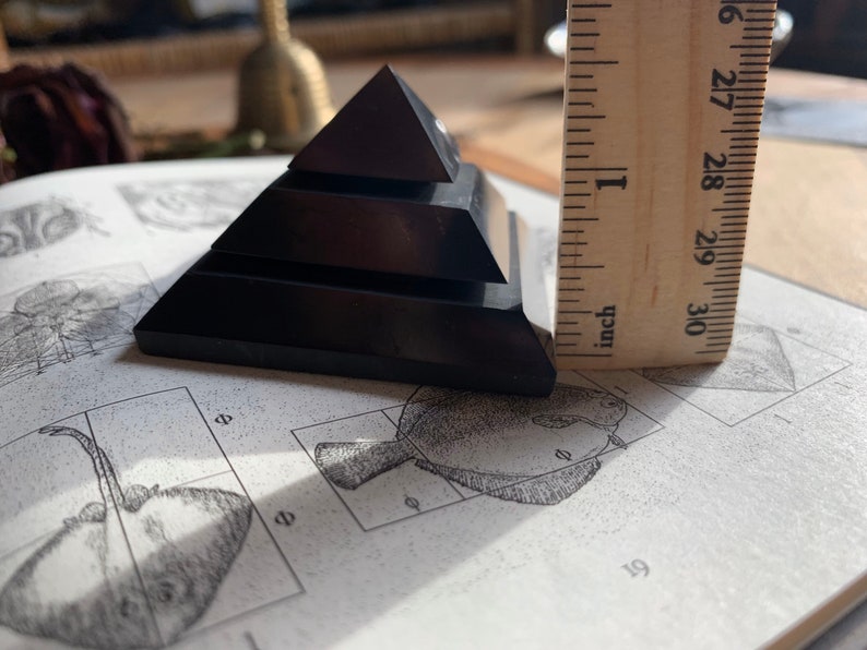May include: A black obsidian pyramid with four tiers, measuring approximately 1 inch in height. The pyramid is resting on a page of a book with illustrations.