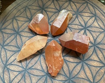 May include: Five orange crystal points arranged in a star shape on a wooden plate with a flower of life design.