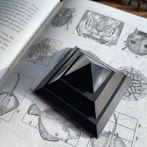 May include: A black obsidian pyramid with a layered design sits on a page of a book with illustrations of animals and geometric shapes. The book page has the text "&Phi;" and "19" printed on it.