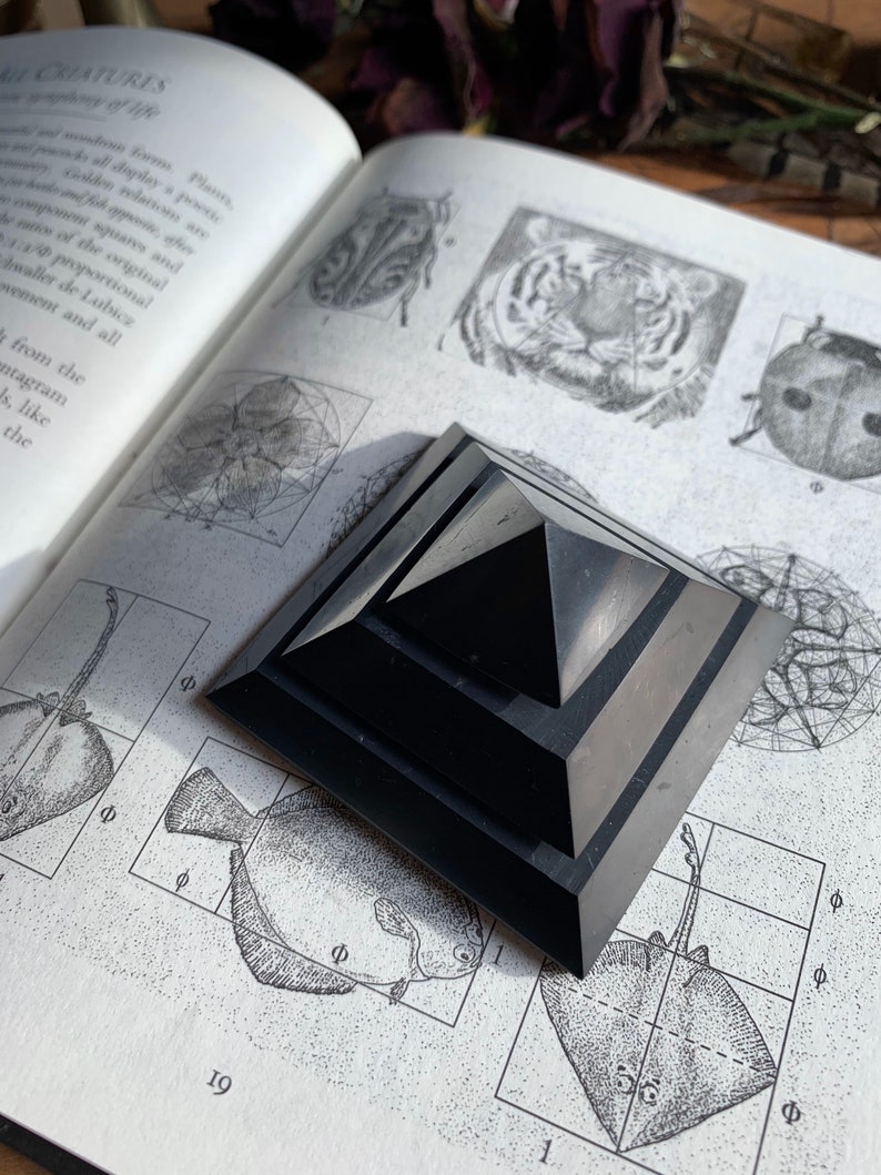 May include: A black obsidian pyramid with a layered design sits on a page of a book with illustrations of animals and geometric shapes. The book page has the text "&Phi;" and "19" printed on it.
