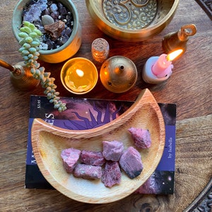 May include: A wooden tray with a variety of items, including a small succulent plant, a brass singing bowl, candles, and a wooden bowl filled with pink crystals. A book titled "Mysteries" is also present.