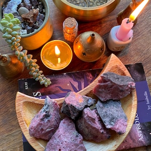 May include: A wooden crescent moon-shaped dish filled with pink and black rhodochrosite crystals. The dish is on top of a book with the title "ASTROLOGY by Isabella" visible.