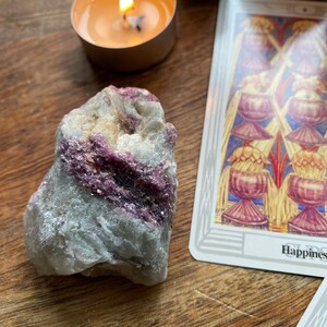 May include: A large, rough, grey and white crystal with streaks of pink and purple. The crystal is sitting on a wooden surface with a lit candle and a tarot card in the background. The tarot card is the "Happiness" card.
