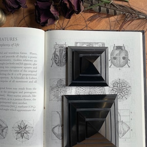 May include: Two black pyramid-shaped objects with multiple layers, sitting on a page of a book with text about geometric forms and the golden ratio. The book is open and the pages are white.