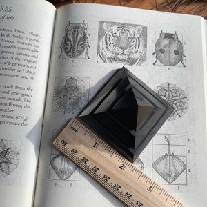 May include: A black obsidian pyramid, approximately 2 inches tall, resting on a page of a book with illustrations of geometric shapes and the golden ratio. The book is open and a wooden ruler is placed on top of the pyramid.
