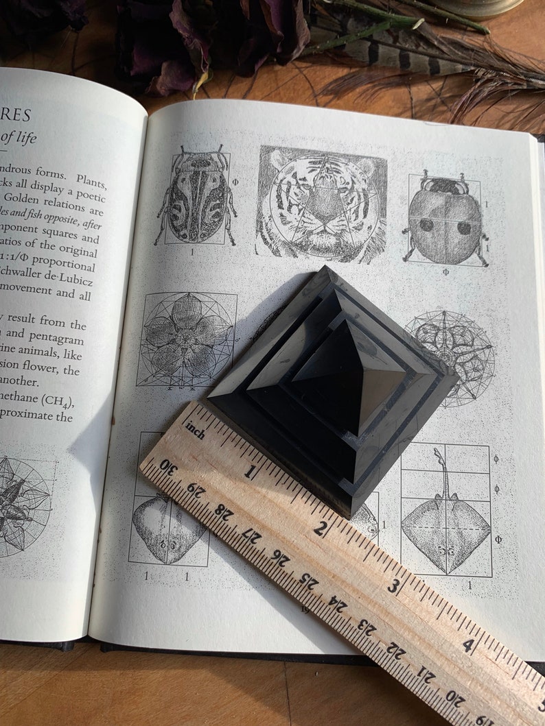 May include: A black obsidian pyramid, approximately 2 inches tall, resting on a page of a book with illustrations of geometric shapes and the golden ratio. The book is open and a wooden ruler is placed on top of the pyramid.