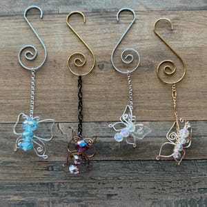 May include: Four decorative ornaments with butterfly designs. Each ornament features a wire butterfly with beaded accents, suspended from an S-shaped hook. The hooks are silver and gold-toned, with a decorative swirl.