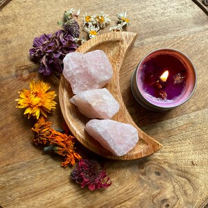 May include: A wooden crescent-shaped dish holds three raw, light pink rose quartz crystals. A lit candle in a silver tin with a purple wax and dried flowers sits nearby. Dried flowers in shades of yellow, orange, and purple surround the objects.