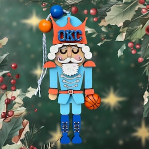 Puede incluir: Un adorno de cascanueces de madera azul y naranja con un balón de baloncesto en la mano. El cascanueces lleva una corona con las letras "OKC" en ella.