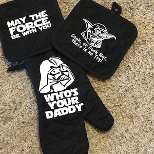 May include: Set of three black Star Wars themed oven mitts. One mitt features Darth Vader with the text "Who's Your Daddy". Another mitt features Yoda with the text "Cook, or Cook Not, there is no Try!" The third mitt features the text "May the Force Be With You".